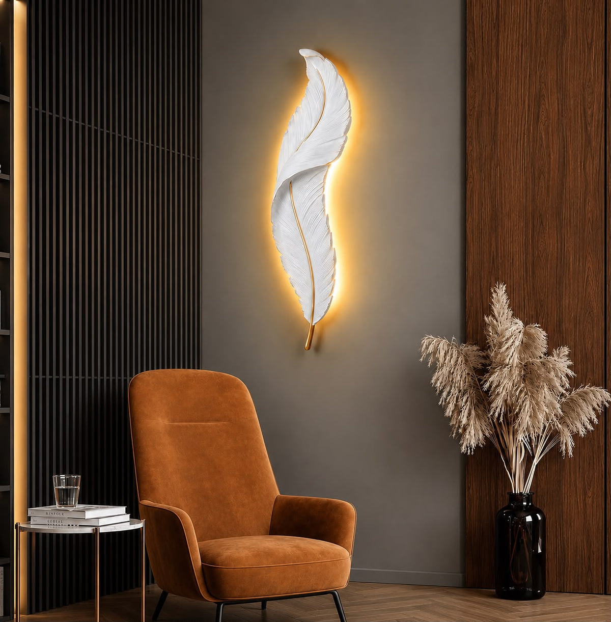 A modern feather-shaped wall sconce with a white textured feather design and a warm yellow glow, mounted on a dark gray wall in a styled interior next to an orange armchair and a vase of pampas grass.